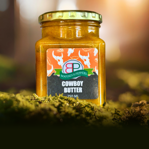 A square jar of Booysens Peppers Cowboy Butter on green moss, with a blurred forest and sunlight in the background.