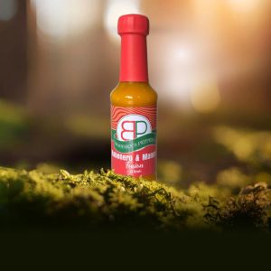 A bottle of Booysens Peppers Habanero & Mango Fusion hot sauce on green moss, with a blurred forest and sunlight in the background.
