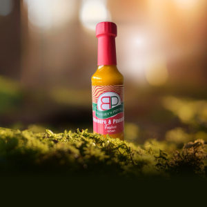 A bottle of Booysens Peppers Habanero & Pineapple Fusion hot sauce on green moss, with a blurred forest and sunlight in the background.