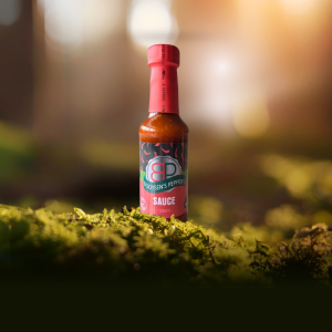 A bottle of Booysens's Peppers Sauce stands on a bed of green moss. The red and black label with a chili pepper logo is visible, and the bottle size is marked as 125ml. The background is a blurred forest scene with a warm, sunlit glow.