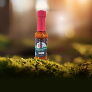 A small glass bottle of "Booysens Peppers" sauce stands on a vibrant green bed of moss. The bottle has a red wax seal and is labeled with the product name and a logo featuring red chilli peppers. The background is a soft, sun-drenched blur of browns and yellows.