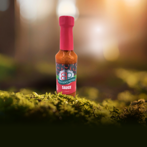 A bottle of Booysen's Peppers Mild Sauce with a red and black label stands on green moss in a sunlit forest.