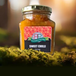 A 250 mL square glass jar of Booysen's Peppers Sweet Chilli Sauce sits on green moss. The sauce is golden-orange with pepper flakes. The label is black and red with a 'BP' logo. Warm, golden sunlight illuminates the jar from a blurred background.