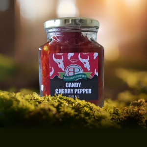 A 250 mL square glass jar of Booysen's Peppers Candy Cherry Pepper sits on bright green moss. The contents are deep red. The label features a black background and a red pattern with white bull skulls.